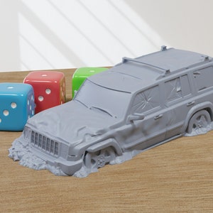 Könnte beinhalten: Ein graues Spielzeugauto mit einer zerbrochenen Windschutzscheibe und einem Dachgepäckträger. Das Auto ist teilweise in grauem Sand versunken. In der Nähe befinden sich drei bunte Würfel, einer blau, einer rot und einer grün.