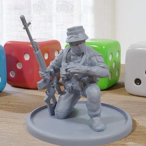May include: A gray plastic miniature figurine of a soldier kneeling with a rifle, wearing a hat and a face mask. The figurine is on a gray plastic base. There are four dice in the background, two blue, one red, and one green.