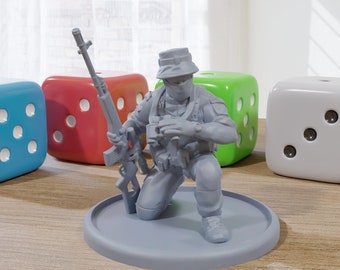 May include: A gray plastic miniature figurine of a soldier kneeling with a rifle, wearing a hat and a face mask. The figurine is on a gray plastic base. There are four dice in the background, two blue, one red, and one green.