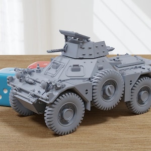 Op de afbeelding: Een grijs model van een gepantserd voertuig met zes wielen, een geschutskoepel en een machinegeweer. Een blauwe dobbelsteen en een rode dobbelsteen liggen naast het voertuig. Het model staat op een houten oppervlak.