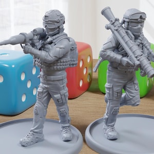 May include: Two gray miniature soldier figurines, each holding a weapon, stand on gray circular bases. The figures are detailed with tactical gear. A blue, red, green, and white die are in the background. The scene is set on a wooden surface.