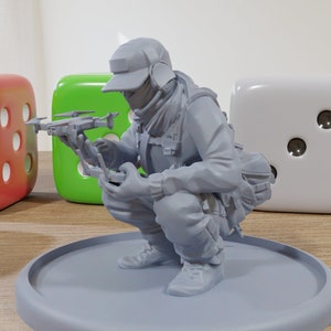 May include: A gray figurine of a person wearing a gray hat and scarf, holding a small gray drone in their hands. The figurine is sitting on a gray circular base.