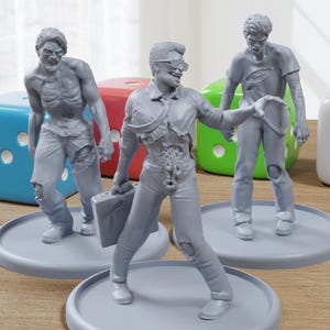 Op de afbeelding: Vier grijs geverfde miniatuurfiguren van zombies, elk staand op een grijze basis. De figuren zijn in verschillende posities geposeerd, met één die een aktetas vasthoudt. De figuren staan op een houten oppervlak met drie dobbelstenen op de achtergrond.