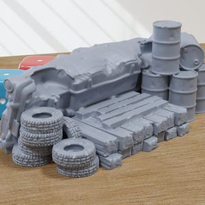 Puede incluir: Un modelo de coche destrozado gris con una pila de tablones de madera y varios barriles grises. El coche destrozado está rodeado por una pila de neumáticos.