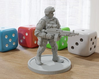 May include: A gray plastic miniature figurine of a soldier holding a machine gun. The soldier is wearing a helmet and tactical gear. The figurine is standing on a gray base. There are three dice in the background, one blue, one red, and one white.