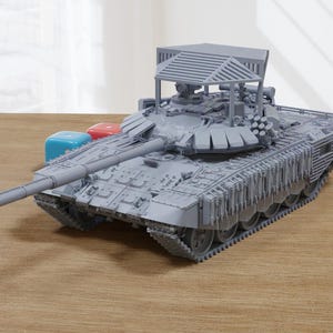 Op de afbeelding: Een grijs plastic model van een tank met een gedetailleerd ontwerp. De tank heeft een groot kanon en een beschermende kooi bovenop. Het model staat op een houten oppervlak.