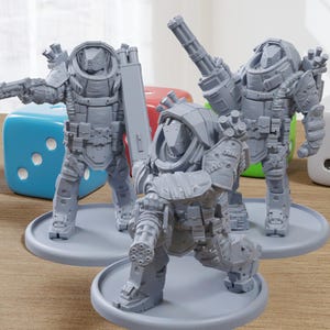 May include: Three gray miniature figurines of futuristic soldiers, each posed with weapons and armor. The figures are on gray circular bases, with a blue, red, and green die and a white die in the background. The figurines are on a wooden surface.