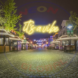 May include: A yellow neon sign with the text "it's a vibe" hanging above a cobblestone street lined with shops and restaurants at night.