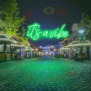 May include: A green neon sign with the text "it's a vibe" hanging above a cobblestone street at night. The sign is illuminated and the street is lined with small shops and businesses.