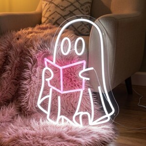 May include: A neon sign shaped like a ghost reading a book. The ghost outline is illuminated in bright white light, while the open book held in its hands is outlined in a vibrant pink neon light. The sign is mounted on a clear acrylic backing.
