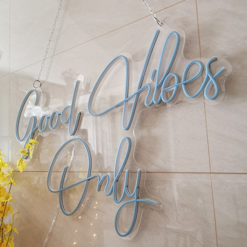 May include: A blue neon sign with the words "Good Vibes Only" hanging on a white tiled wall. The sign is made of glass and has a clear acrylic backing.