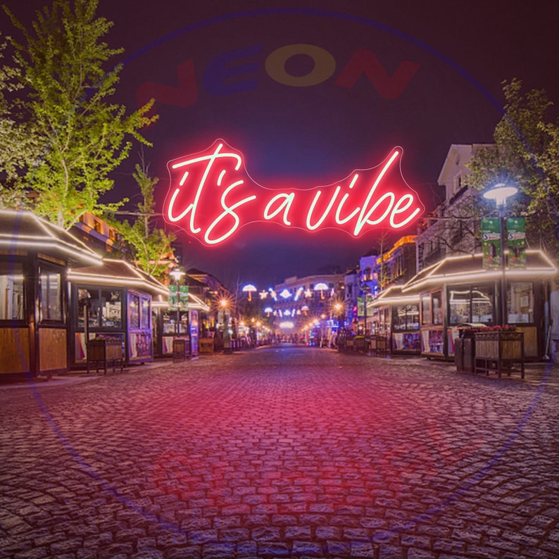 May include: A red neon sign with the text "it's a vibe" hanging above a cobblestone street lined with small shops at night.