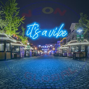 May include: A blue neon sign with the text "it's a vibe" in a cursive font. The sign is hanging above a cobblestone street lined with shops and restaurants at night.