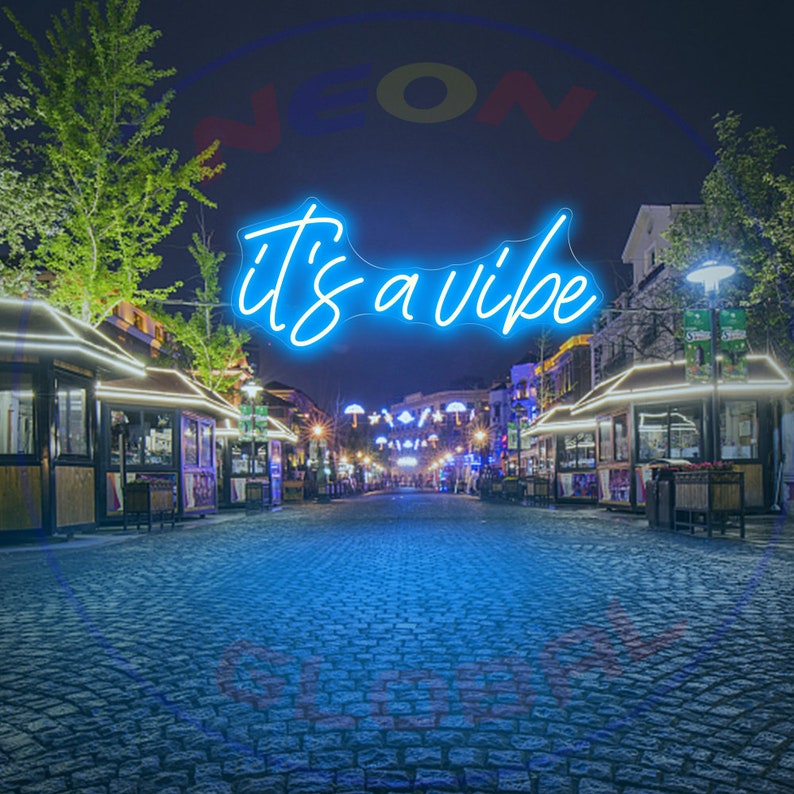 May include: A blue neon sign with the text "it's a vibe" in a cursive font. The sign is hanging above a cobblestone street lined with shops and restaurants at night.
