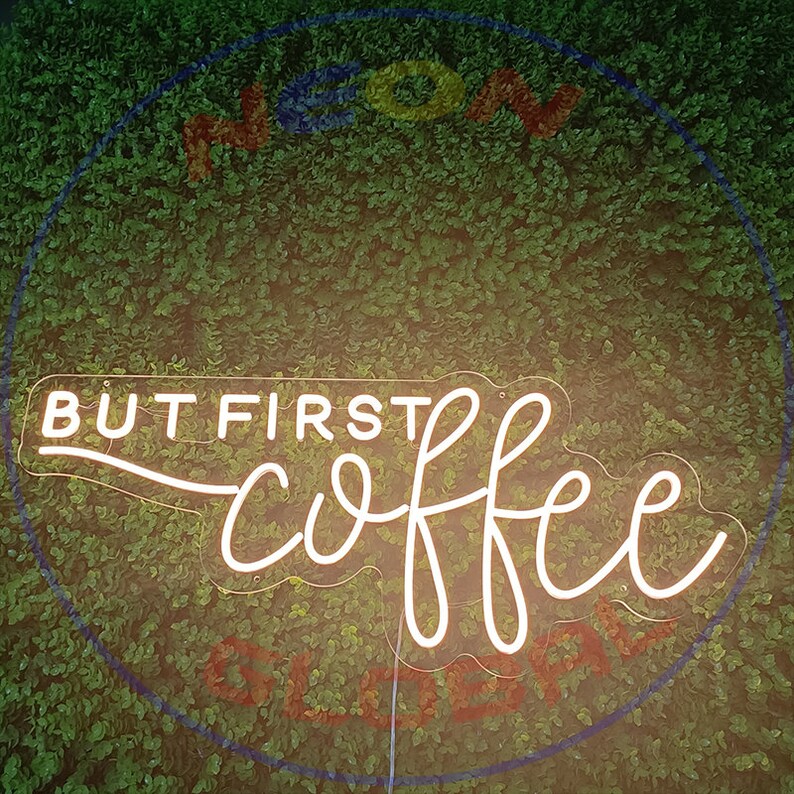 May include: White neon sign with the text "But first, coffee" in a cursive font. The sign is mounted on a green wall.