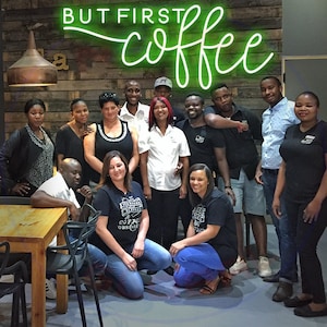 May include: A group of people stand and sit in front of a wooden wall with a neon sign that reads "But First Coffee". The people are smiling and looking at the camera. Some are wearing black shirts with the words "Coffee Obsession" printed on them.