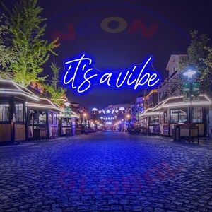 May include: Blue neon sign with the text "it's a vibe" hanging above a cobblestone street at night. The sign is illuminated and the street is lined with shops and restaurants.
