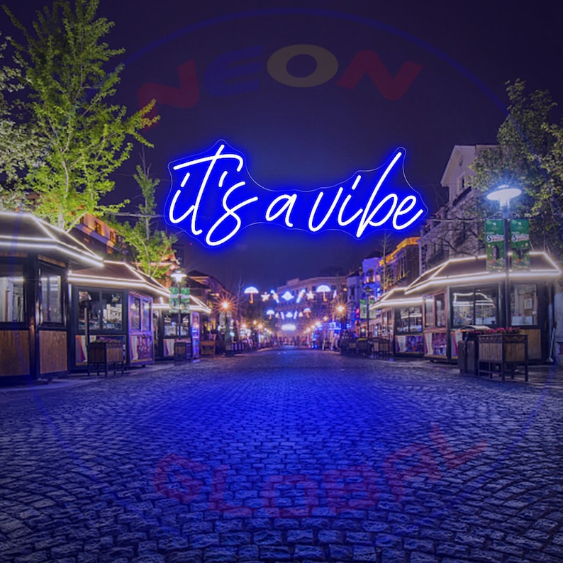 May include: Blue neon sign with the text "it's a vibe" hanging above a cobblestone street at night. The sign is illuminated and the street is lined with shops and restaurants.