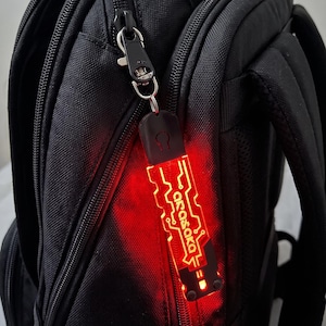 Pode incluir: Um porta-chaves retangular preto com uma luz vermelha brilhante e um padrão de circuito impresso. O texto "arasaka" está impresso a branco na parte da frente. O porta-chaves está preso ao fecho de uma mochila preta com um mosquetão.