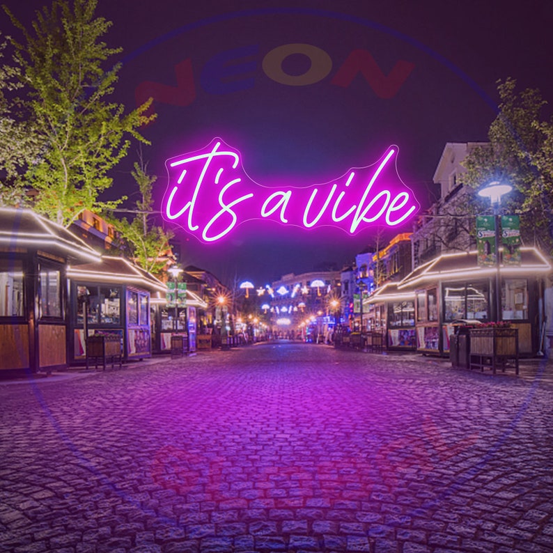 May include: A pink neon sign with the text "it's a vibe" hanging above a cobblestone street at night. The street is lined with small shops and buildings with string lights.