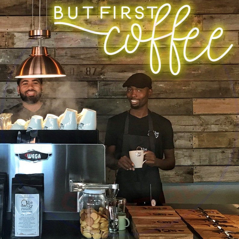 May include: Two people stand behind a coffee bar with a sign that reads "BUT FIRST coffee" in yellow neon lettering. The person on the left is wearing a black shirt and the person on the right is wearing a black shirt with a black apron. The person on the right is holding a white coffee mug.
