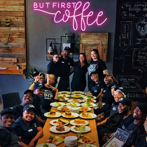 May include: A group of people are gathered around a table with plates of food. The table is covered in a brown tablecloth and there are many plates of food on it. The people are all smiling and laughing. The sign above the table says "BUT FIRST COFFEE".