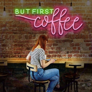 May include: A woman sits at a counter in a cafe, looking at a tablet. A neon sign above her reads "BUT FIRST coffee" in pink and green letters.