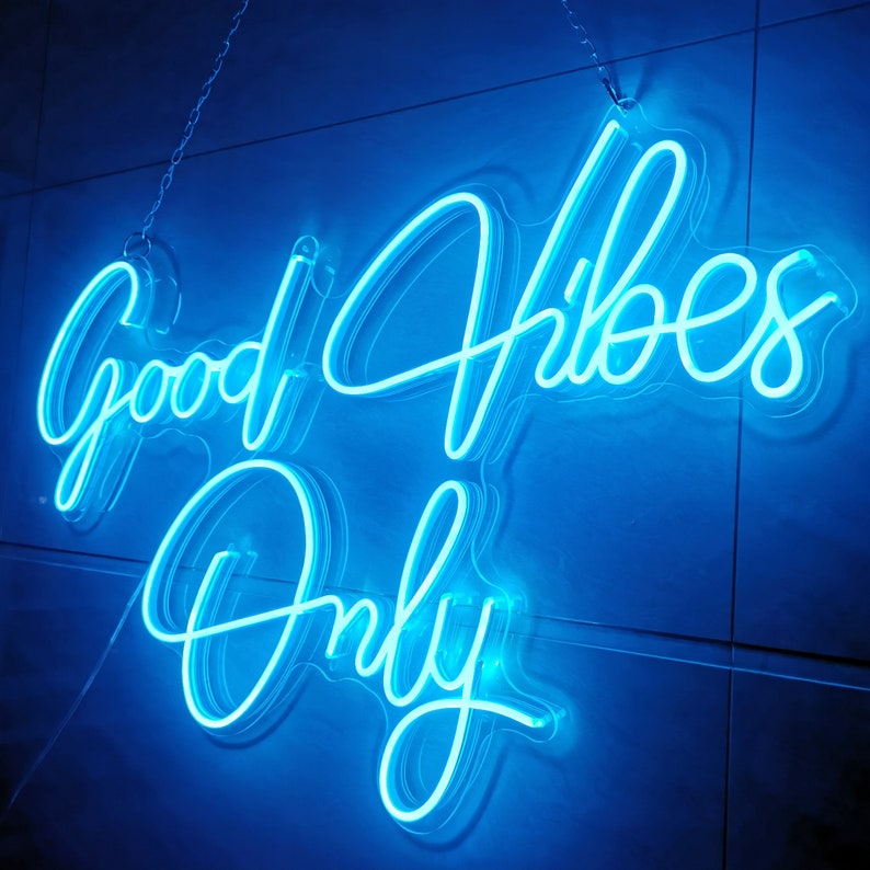 May include: A neon sign with the words "Good Vibes Only" in a bright blue color. The sign is hanging from a chain and is illuminated against a white tiled wall.