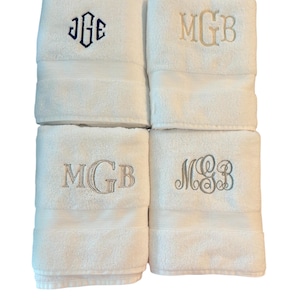 May include: Four white bath towels, each embroidered with initials. Two towels feature "MGB" in light brown, one in dark blue, and one in light grey. Each towel has a decorative stripe near the bottom.