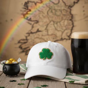 Op de afbeelding: Een witte baseballcap met een groene klaverbladpatch en gouden rand. Een zwarte pot met goud, een pint donker bier en klaverbladconfetti liggen op een houten oppervlak. Een kaart en een regenboog zijn op de achtergrond.