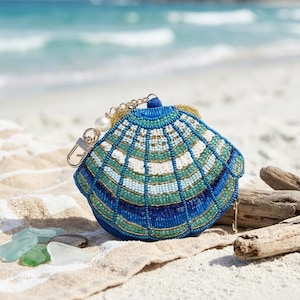 Könnte beinhalten: Eine muschelförmige Clutch mit Perlen in Blau-, Weiß- und Goldtönen. Sie hat eine goldene Kette und eine Perle. Sie liegt auf einem Sandstrand, mit dem Meer im Hintergrund.