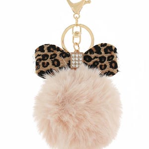 Könnte beinhalten: Ein Schlüsselanhänger mit einem rosa Kunstfell-Pompon, einer Schleife mit Leopardenmuster und goldfarbenen Beschlägen. Die Schleife hat ein braunes und schwarzes Fleckenmuster. Ein kleiner, rechteckiger Anhänger mit klaren Strasssteinen ist befestigt.
