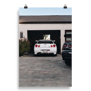 May include: A white sports car with red taillights is parked in a garage. A black sports car is parked next to it. The cars are on a paved driveway in front of a house.