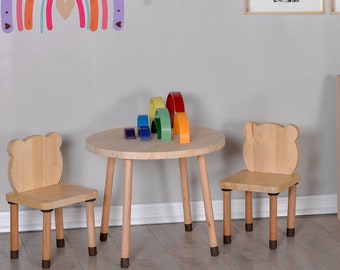 May include: A children's playroom scene featuring a small wooden table and two chairs with bear-shaped backs. A rainbow-shaped wall decal and a wooden rainbow stacking toy sit on the table. A bookshelf and framed animal prints complete the scene.