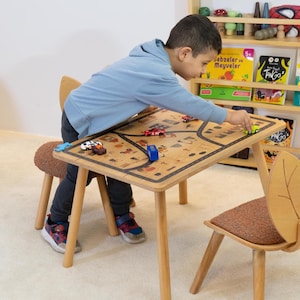 May include: A child plays with toy cars on a wooden table with a road map design. The table has two wooden chairs, one with a brown cushion and the other with a brown and orange cushion. A bookshelf with books and toys is in the background.