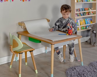 May include: A wooden children's art table with a roll of paper and a green storage bin. A child sits at the table with a cup of crayons and a pencil. The table has light blue accents on the legs.