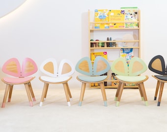 Puede incluir: Cinco sillas infantiles coloridas con un diseño único de respaldo en forma de mariposa. Las sillas tienen patas de madera y asientos en rosa, blanco, azul, verde y gris. Las sillas están dispuestas en fila en una sala de juegos moderna y luminosa.