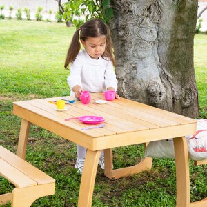 May include: A light brown wooden picnic table with a matching bench is set up on a grassy lawn. A young child is playing with a pink tea set, including cups, saucers, and a small pitcher. The scene is outdoors, near a large tree.