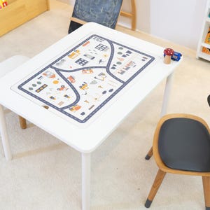Puede incluir: Una mesa blanca con una alfombra de juego que presenta un diseño de mapa de carreteras de la ciudad. La alfombra de juego tiene un borde azul y presenta pequeños coches, edificios y árboles. Una pequeña silla de madera con un asiento negro y orejas de conejo está al lado de la mesa.