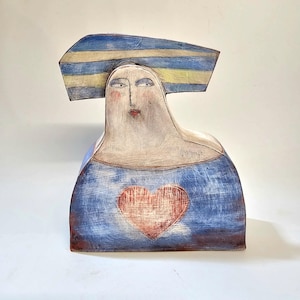 May include: A ceramic sculpture of a woman with a blue and yellow striped hat, wearing a blue dress with a red heart on the front.