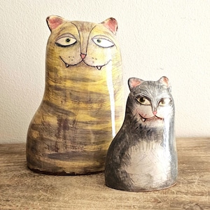 May include: Two ceramic cat figurines with a glossy finish. The larger cat is yellow with brown tabby stripes, while the smaller cat is grey with white accents. Both figures feature hand-painted faces with green eyes, pink noses, and small fangs.