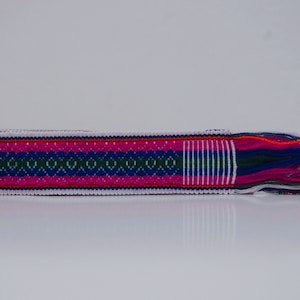 Può includere: Una cintura bianca tessuta con un motivo geometrico rosa, blu e verde. La cintura ha lunghe frange di diversi colori.