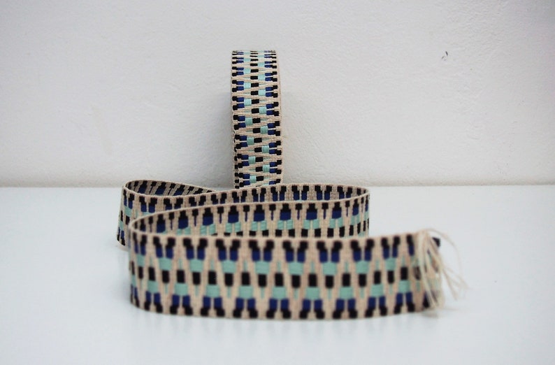 May include: A close-up of a beige, blue, and black patterned ribbon. The ribbon is woven with a geometric design.