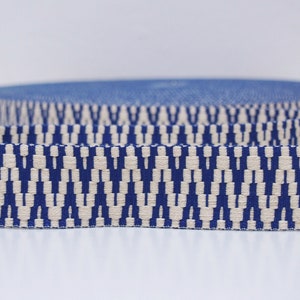 May include: A roll of blue and cream-colored woven ribbon. The ribbon has a repeating geometric pattern in a dark blue color against a cream background. The ribbon is likely used for crafting, sewing, or decorative purposes.