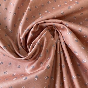 Könnte beinhalten: Nahaufnahme eines staubrosafarbenen Stoffes mit zartem Blumenmuster. Der Stoff ist mit kleinen, verstreuten Blumen in Weiß- und Blautönen verziert. Das Material ist drapiert und erzeugt weiche Falten und Schatten.