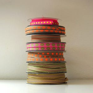 Op de afbeelding: Een stapel decoratieve lintrollen in verschillende kleuren. De linten omvatten bruin met oranje bloempatronen, roze met groene bloempatronen en bruin met oranje stippen.