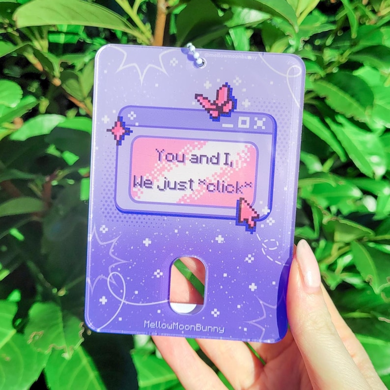 Puede incluir: Encanto de acr&iacute;lico morado y rosa con un dise&ntilde;o pixelado de una ventana de computadora con el texto "You and I, We just "click"" y una mariposa rosa. El encanto tiene un agujero en la parte superior para colgar.