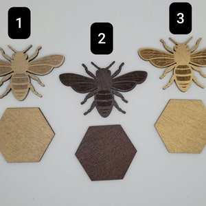 Puede incluir: Tres recortes de abejas de madera, cada uno con una mancha de madera diferente. Las abejas est&aacute;n numeradas del 1 al 3. Cada abeja est&aacute; sentada sobre un recorte hexagonal de madera.