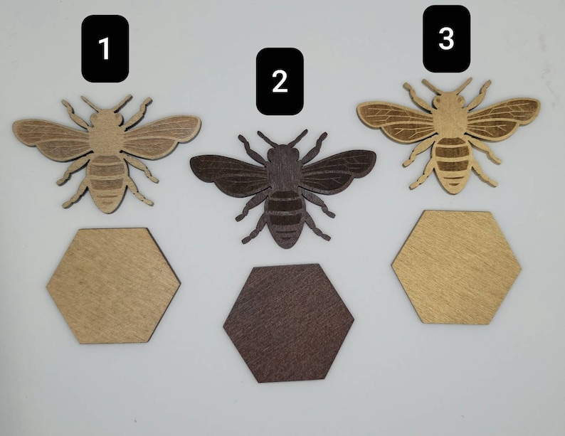Puede incluir: Tres recortes de abejas de madera, cada uno con una mancha de madera diferente. Las abejas est&aacute;n numeradas del 1 al 3. Cada abeja est&aacute; sentada sobre un recorte hexagonal de madera.