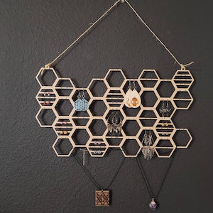Puede incluir: Organizador de pendientes y collares de madera en forma de panal con m&uacute;ltiples compartimentos. El organizador est&aacute; colgado de una cuerda y tiene varios pendientes y collares colgando de &eacute;l.